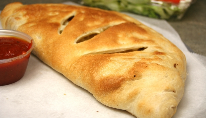 Golden-brown calzone on parchment paper with a cup of marinara sauce on the side.