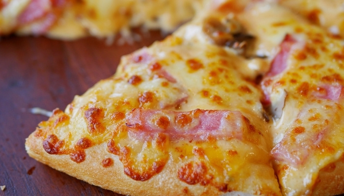 A close-up of a cheesy pizza slice with pieces of ham on a wooden surface.