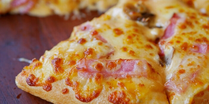 A close-up of a cheesy pizza slice with pieces of ham on a wooden surface.